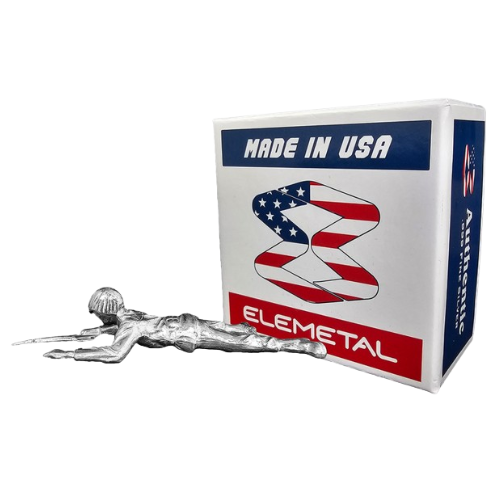 A sculpted silver figure lies next to a white box. The box has "MADE IN USA" at the top, a stylized American flag graphic in the middle, and "ELEMETAL" at the bottom.